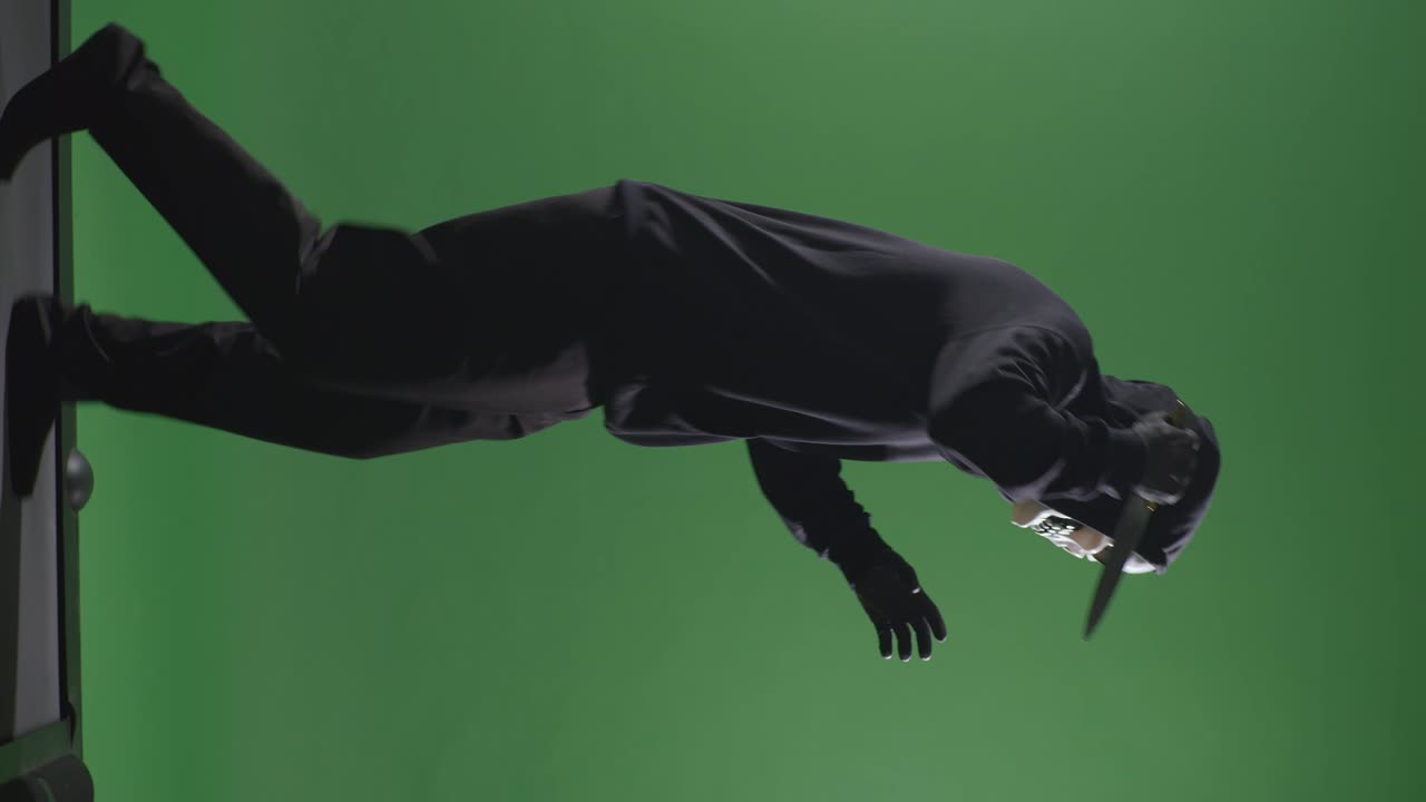 녹색 스크린 배경에서 달리는 동안 칼을 들고 카메라를 가리키는 할로윈 마스크를 입은 후드 스웨트 셔츠를 입은 무서운 남자의 측면 시각