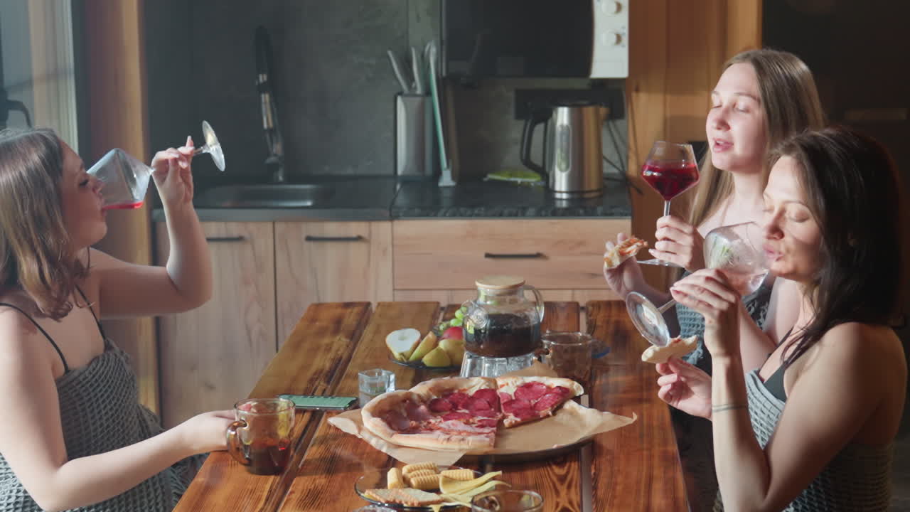 three women around wooden table making toast with wine glasses while enjoying pizza and fruit platter in cozy kitchen setting, two glasses nearly empty and one glass still full, sunlight