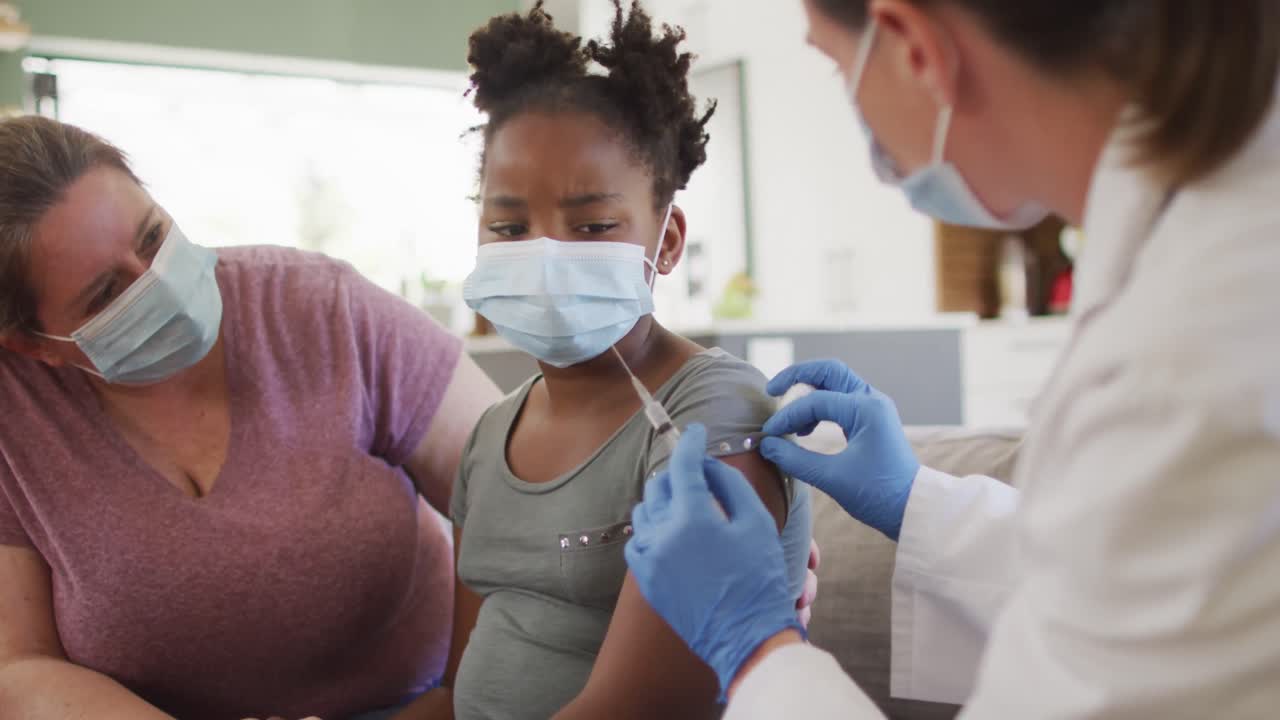 African american girl with caucasian mother and female doctor wearing face masks, vaccinating