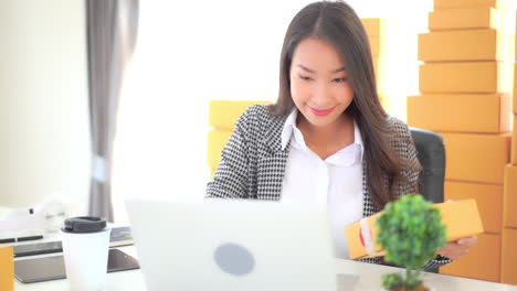 asian business woman entrepreneur arranging orders while sitting at the work desk in front of laptop with yellow parcel in hand, yellow boxes on background