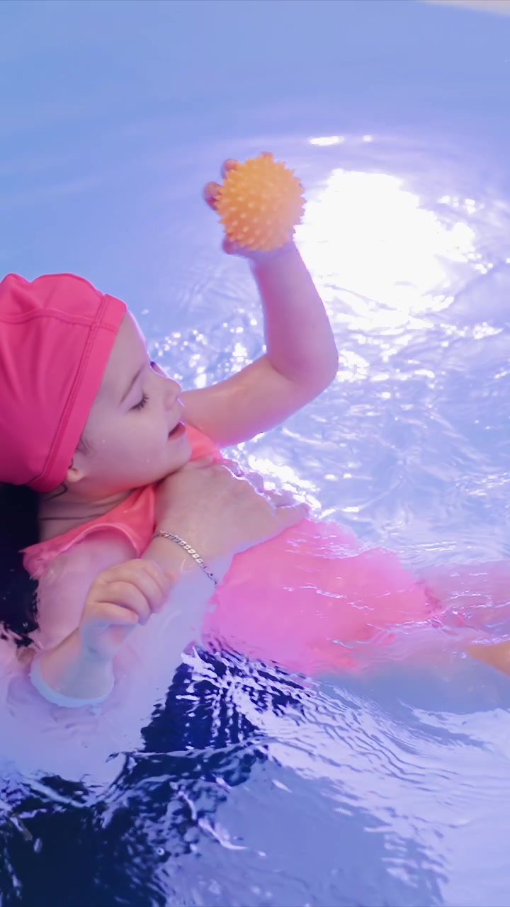 Child is held up while floating surrounded by pool toys. Supportive grip around chest and back keeps baby steady. Blue edge is visible near top frame in consistent setting