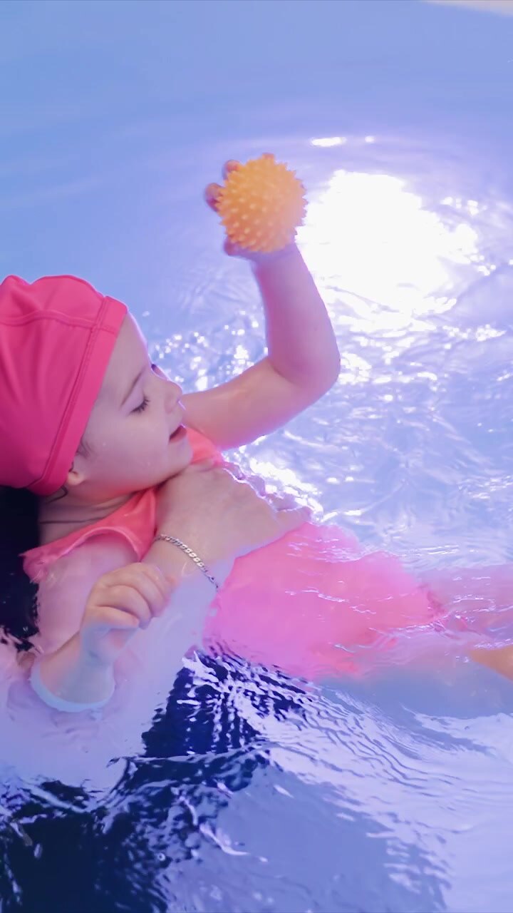 Child is held up while floating surrounded by pool toys. Supportive grip around chest and back keeps baby steady. Blue edge is visible near top frame in consistent setting