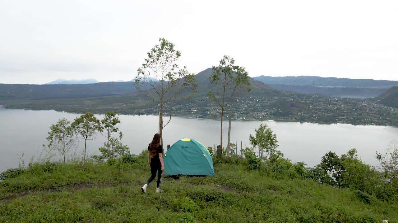 Backpacker girl walking past tent to look at view from a top green hill. Drone fly over.