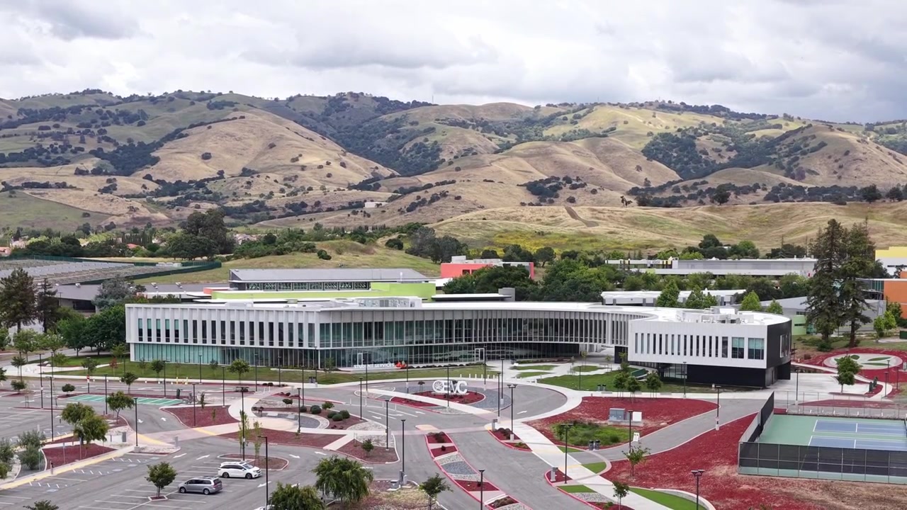 Aerial view descending toward the modern campus of Evergreen Valley College in San Jose, showing the architectural building, empty parking lot, and the beautiful rolling hills of California