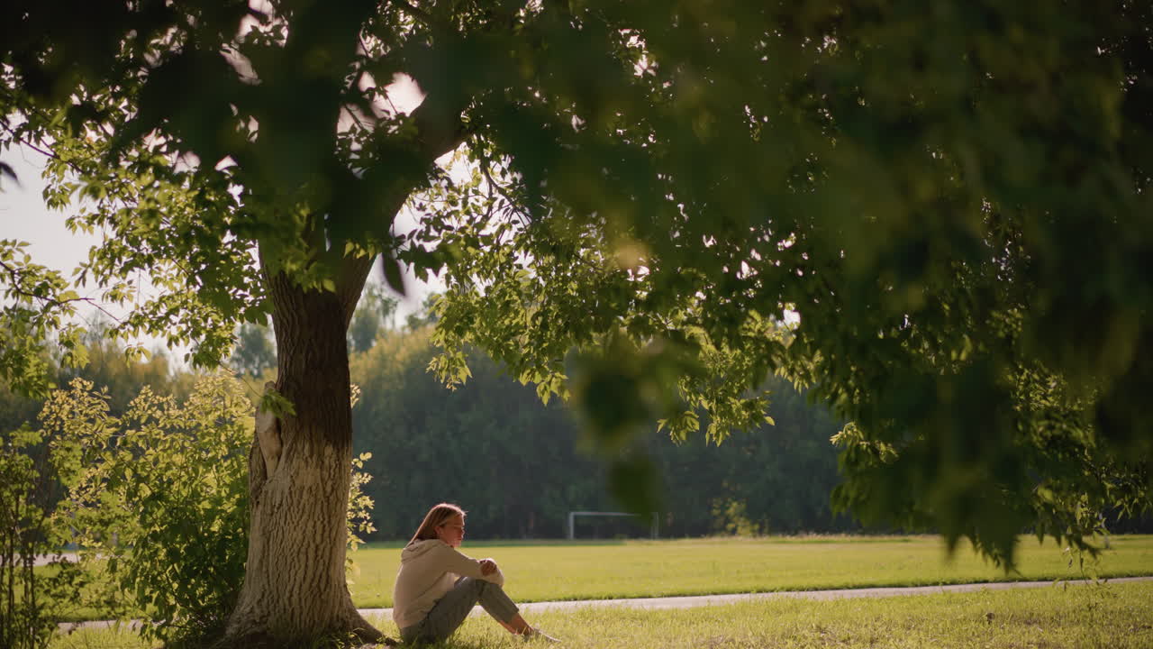 나무 에 앉아 있는 여자, 팔 을 다리 위 에 접어 놓고 사려 깊은 반영 을 하고, 배경 에 골포스트 가 있는 부드러운 초록색 으로 둘러싸여 있다