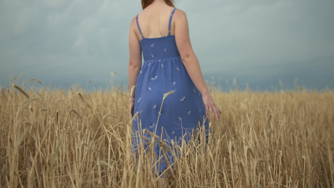 A young woman wearing a blue dress walking in slow motion toward distant dark blue storm clouds.