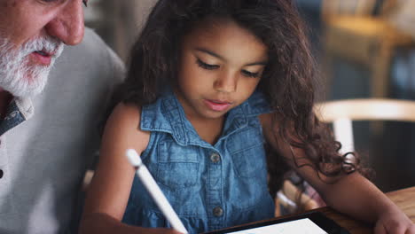 Close-up-of-young-girl-drawing-with-tablet-and-stylus,-with-her-grandfather-sitting-beside-her