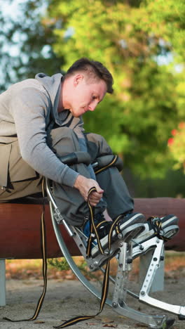 young man with short brown hair sitting on large log in park-like setting, wearing light gray sweatshirt and khaki pants, focusing on adjusting stilt straps with concentration, green trees