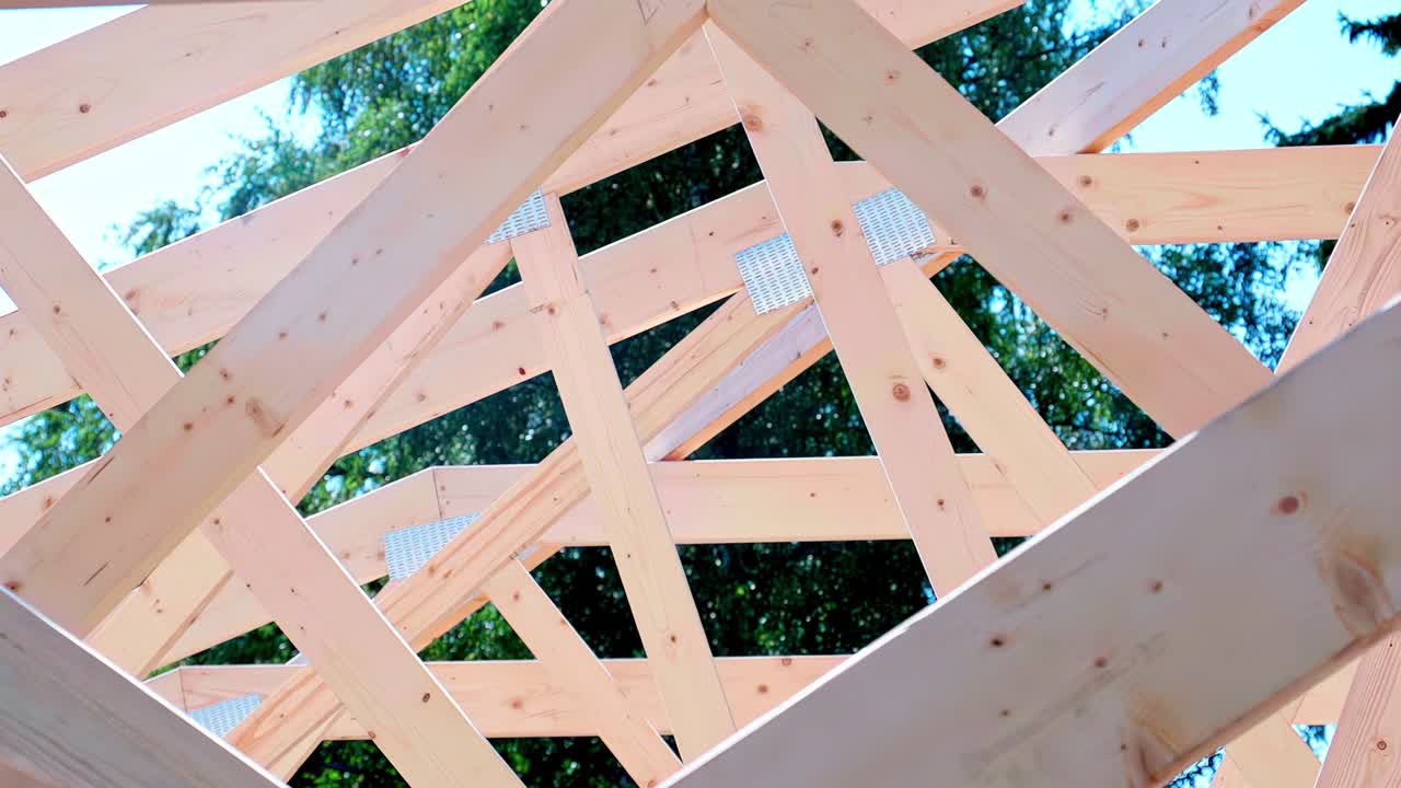 Inquadratura dal basso verso l'alto attraverso le travi in legno della struttura di una casa in costruzione, poi inquadratura dall'alto verso il basso con una leggera panoramica a sinistra e primo piano su un giunto strutturale e un connettore metallico