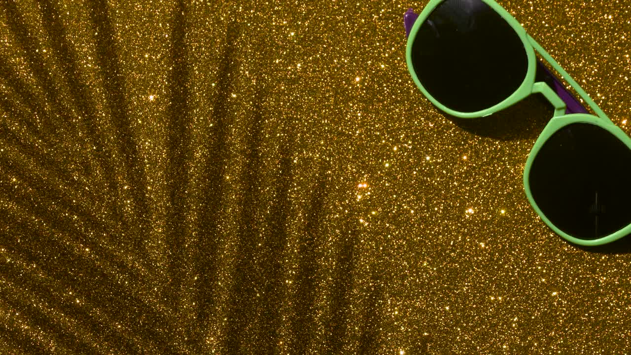 Shadows from a palm tree with sunglasses on a gold glitter sparkling shining background. Minimal art, glamor, vacation, summer, party concept.