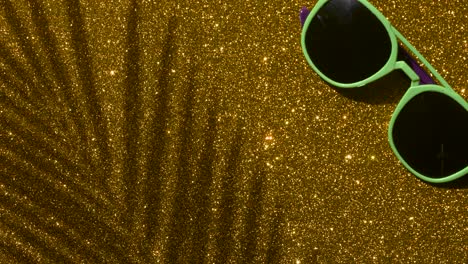 shadows from a palm tree with sunglasses on a gold glitter sparkling shining background. minimal art, glamor, vacation, summer, party concept.