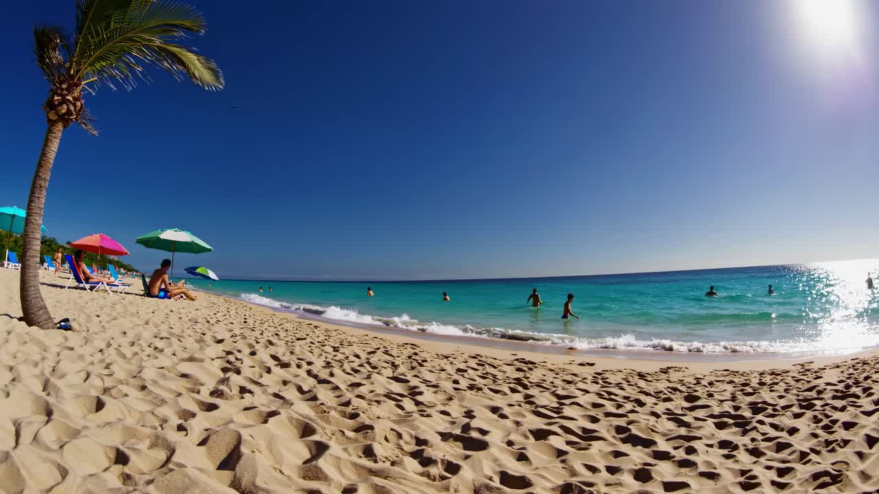 Foto de gran angular de una playa soleada con agua azul cristalina y sombrillas de colores, que captura un ambiente animado