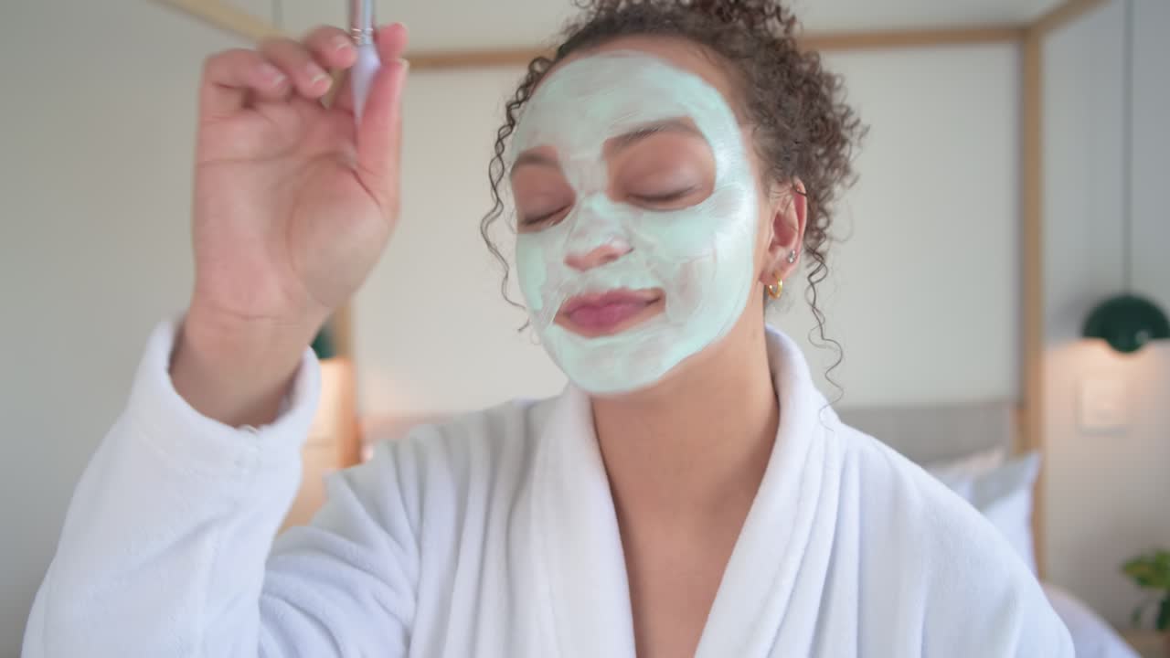 Mujer con bata blanca cogiendo un pincel y un tarro, aplicando una mascarilla verde para el cuidado de la piel junto a una cama con dosel