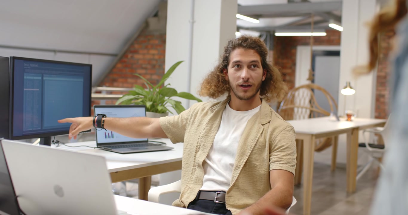 Hombre sentado señalando un monitor y explicando código en una laptop en un loft, mientras un compañero de trabajo entra con un teléfono.