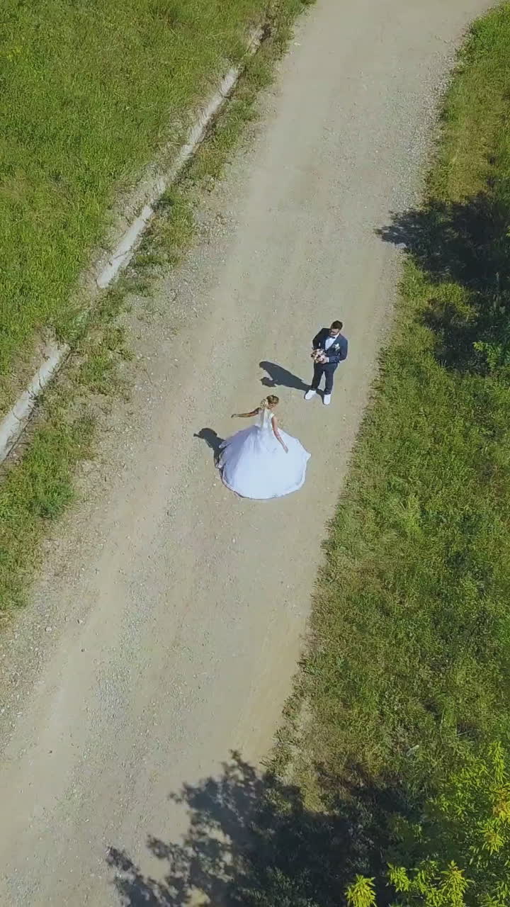 happy young bride spins near watching groom on asphalt road in green park on warm summer day upper view