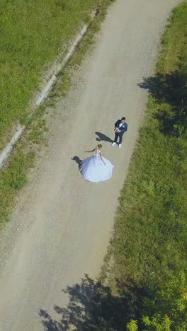 happy young bride spins near watching groom on asphalt road in green park on warm summer day upper view