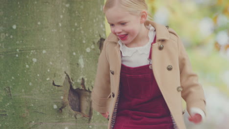 young girl looking around trunk of autumn tree playing hide and seek with father  in garden