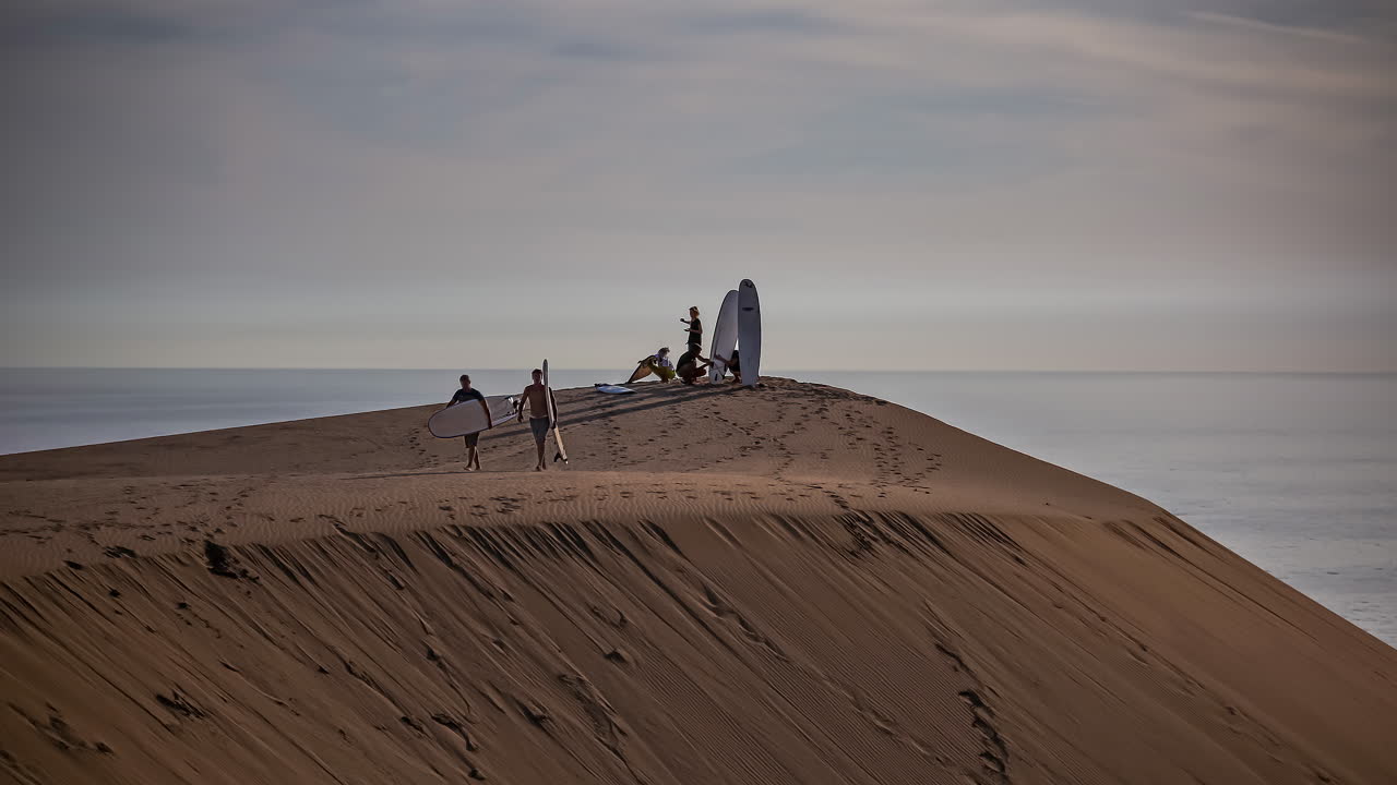 silueta de un grupo de adultos jóvenes surfeando dunas de arena en marruecos con el océano de fondo - lapso de tiempo