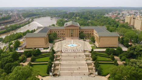 Aerial-drone-view-moving-backward-showing-the-Philadelphia-Museum-of-Art-surrounded-by-bright-green-trees-and-cars-in-the-afternoon-summer-sun