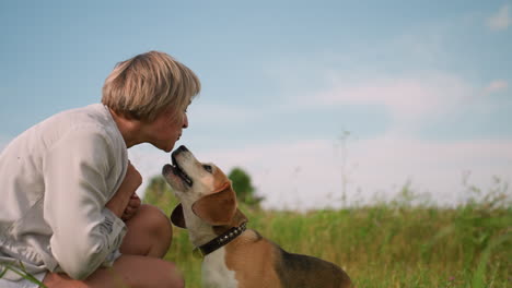 dog owner interacting playfully with her pet as they share a tender moment in outdoor grassy field, owner is squatting down on sunny day while the dog jumps to kiss her