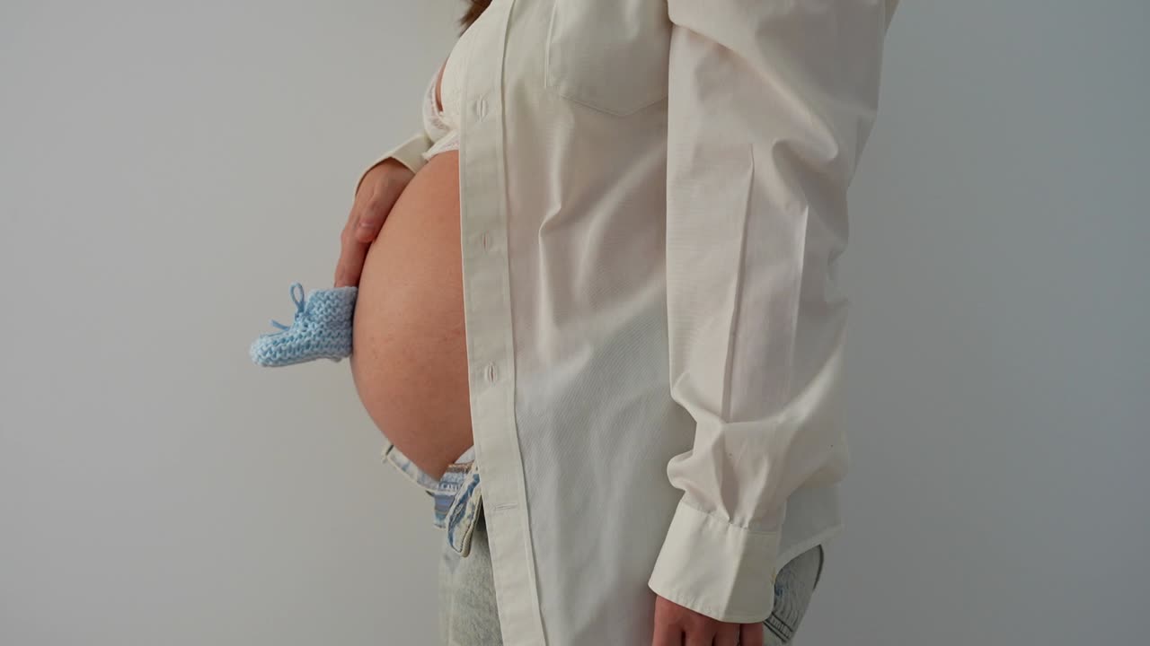 Pancia di una donna incinta che tiene un paio di scarpine blu da neonato. Zoom, sfondo bianco.