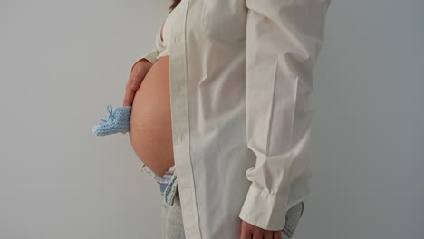 belly of a pregnant woman holding a pair of blue baby boots. zoom in, white background