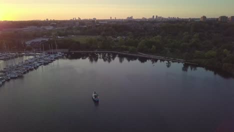 Vista-Aérea-De-La-Puesta-De-Sol-En-órbita-Alrededor-De-Un-Velero-Y-Un-Puerto-Deportivo-En-El-Muelle-Del-Club-De-Yates-En-La-Bahía-Del-Lago-Rodeado-De-árboles-Verdes-Con-El-Horizonte-De-Los-Edificios-De-La-Ciudad-En-El-Fondo-En-Toronto,-Ontario,-Canadá