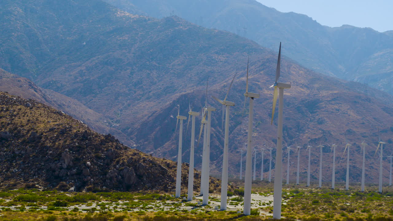 Palm Springs windmills spinning with the rugged San Jacinto Mountains rising behind them in California under a clear blue sky on a bright, sunny day. Aerial shot slowly moving around and away
