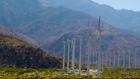 palm springs windmills spinning with the rugged san jacinto mountains rising behind them in california under a clear blue sky on a bright, sunny day. aerial shot slowly moving around and away