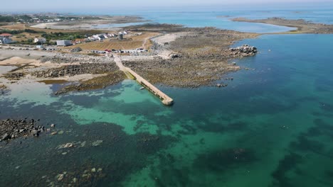 High-circling-drone-footage-of-Guernsey-coastline,-stunning-golden-beach-and-clear-turquoise-calm-sea-with-boats-at-anchor-on-sunny-summer-day