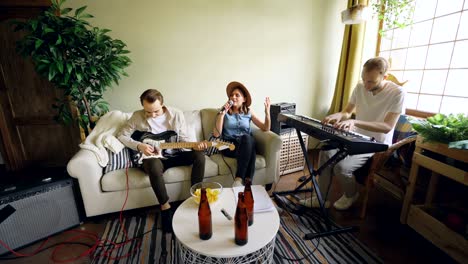 musical band is rehearsing at home, woman is singing and men are playing musical instruments guitar and keyboard. beer bottles and snacks on modern table are visible.