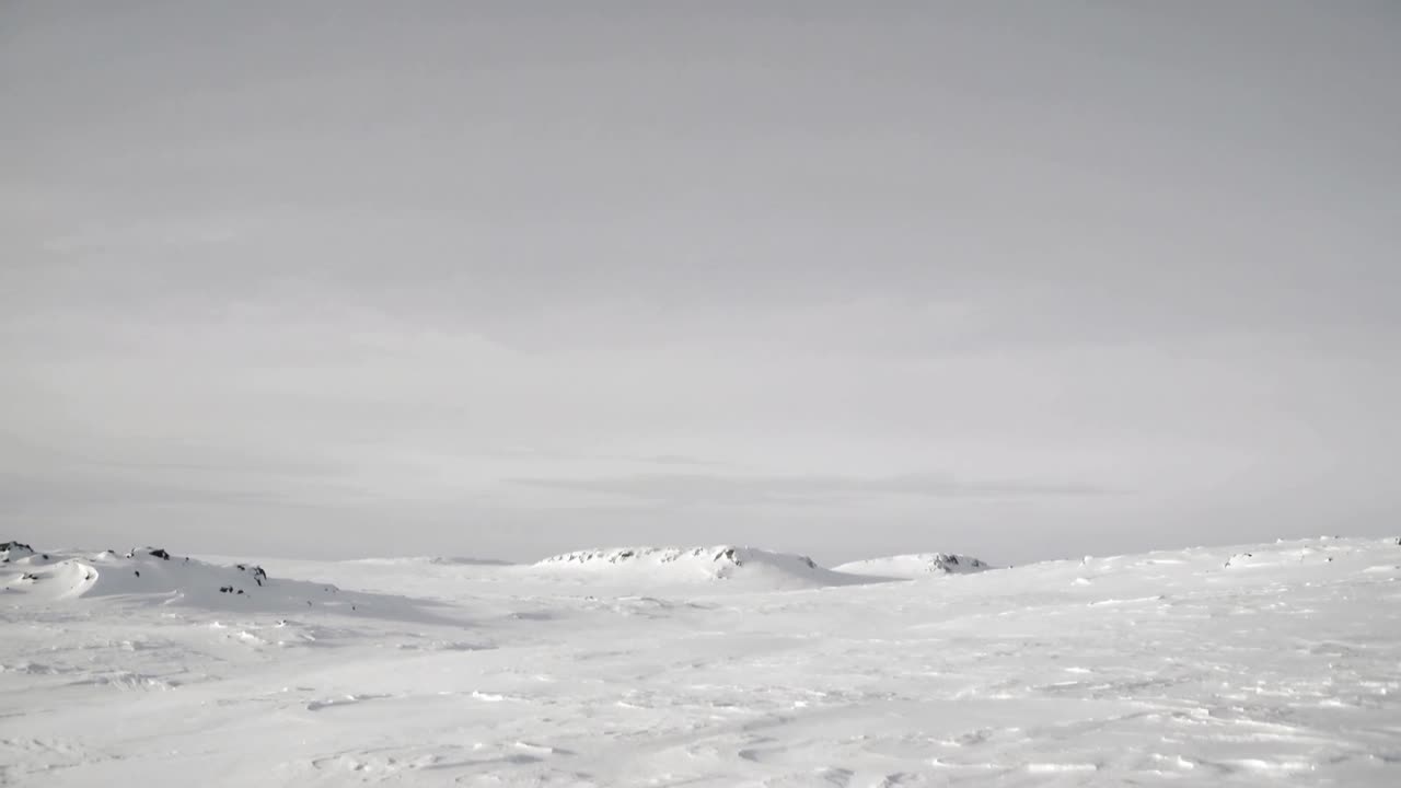 カメラが移動し、曇り空の下の雪原と低い尾根が現れる。表面には波紋が浮かび上がり、その質感を際立たせる。