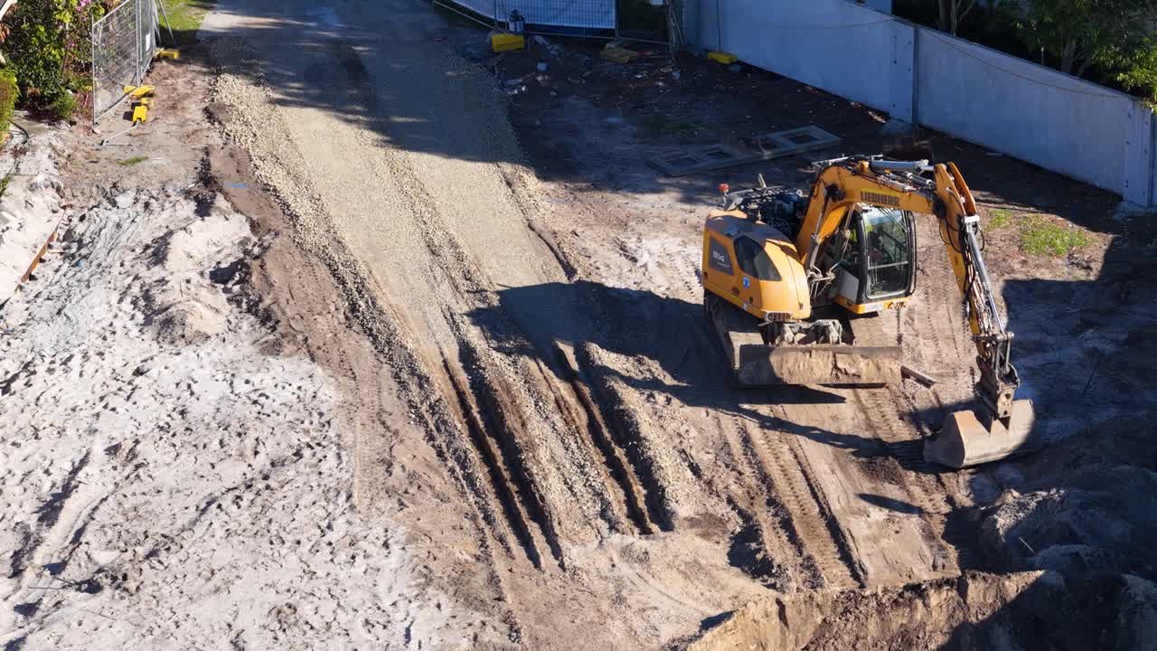 Excavator levels sandy ground at construction site, aerial view, bright daylight, steady camera