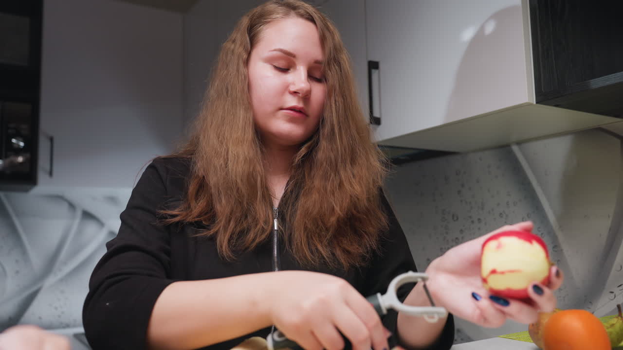 Young lady in modern kitchen peels red apple with handheld peeler, focused expression, blue nails visible, fruit prep under bright light, fresh snack routine, clean countertop background