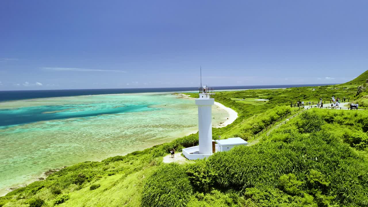 Órbita cinematográfica de drone ao redor do Farol Hirakubozaki branco, revelando uma vasta lagoa de recife de coral turquesa e as águas esmeralda cintilantes da Ilha Ishigaki