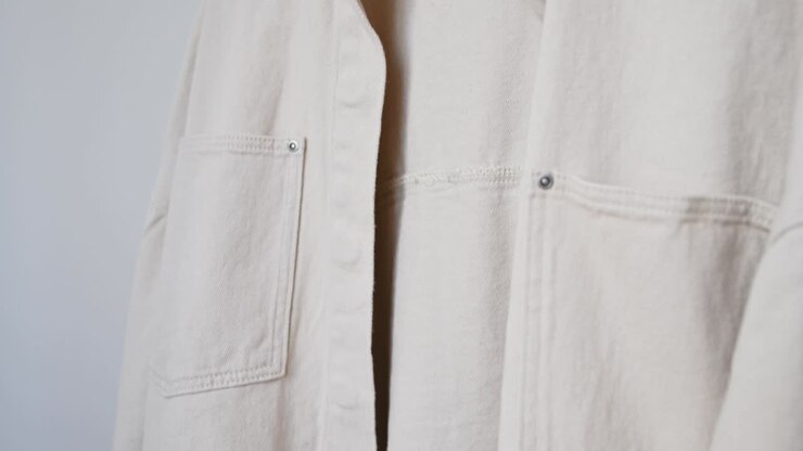 Maglia a maniche lunghe trendy da donna, felpa a righe, camicia di jeans beige, giacca su appendiabiti in legno bianco appesa su stender contro parete bianca. Concetto di abbigliamento minimalista, pubblicità per saldi e shopping in negozio