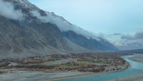 Aerial-scenic-view-of-a-vast-mountain-valley-with-a-river-flowing-through-and-low-clouds-touching-the-mountaintops-in-Nomal-Valley,-Gilgit