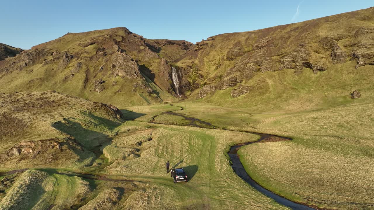 Aerial view of an off-road vehicle parked in a remote valley in northern Iceland, surrounded by grassy hills, winding streams, and a waterfall in the background.