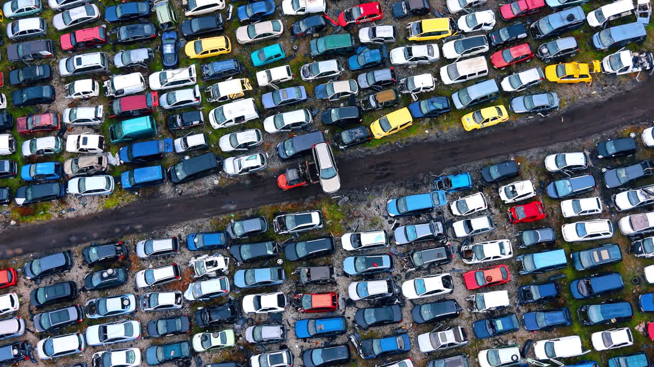 Luftfoto af bilkirkegård med gaffeltruck, der flytter køretøj. Droneoptagelse af organiserede rækker af beskadigede biler og en gaffeltruck, der bærer et køretøj på en genbrugsplads.