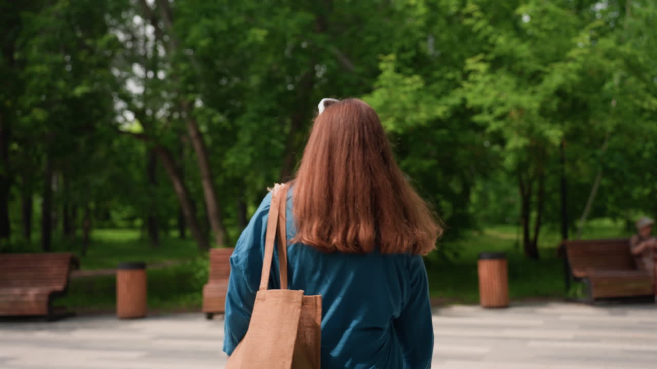 緑豊かな夏の公園を、茶色い長い髪の若い女性が日焼け色のバッグを持って、木のベンチに向かって歩いている後姿。周りには木々が生い茂り、太陽の光が差し込んでいる。