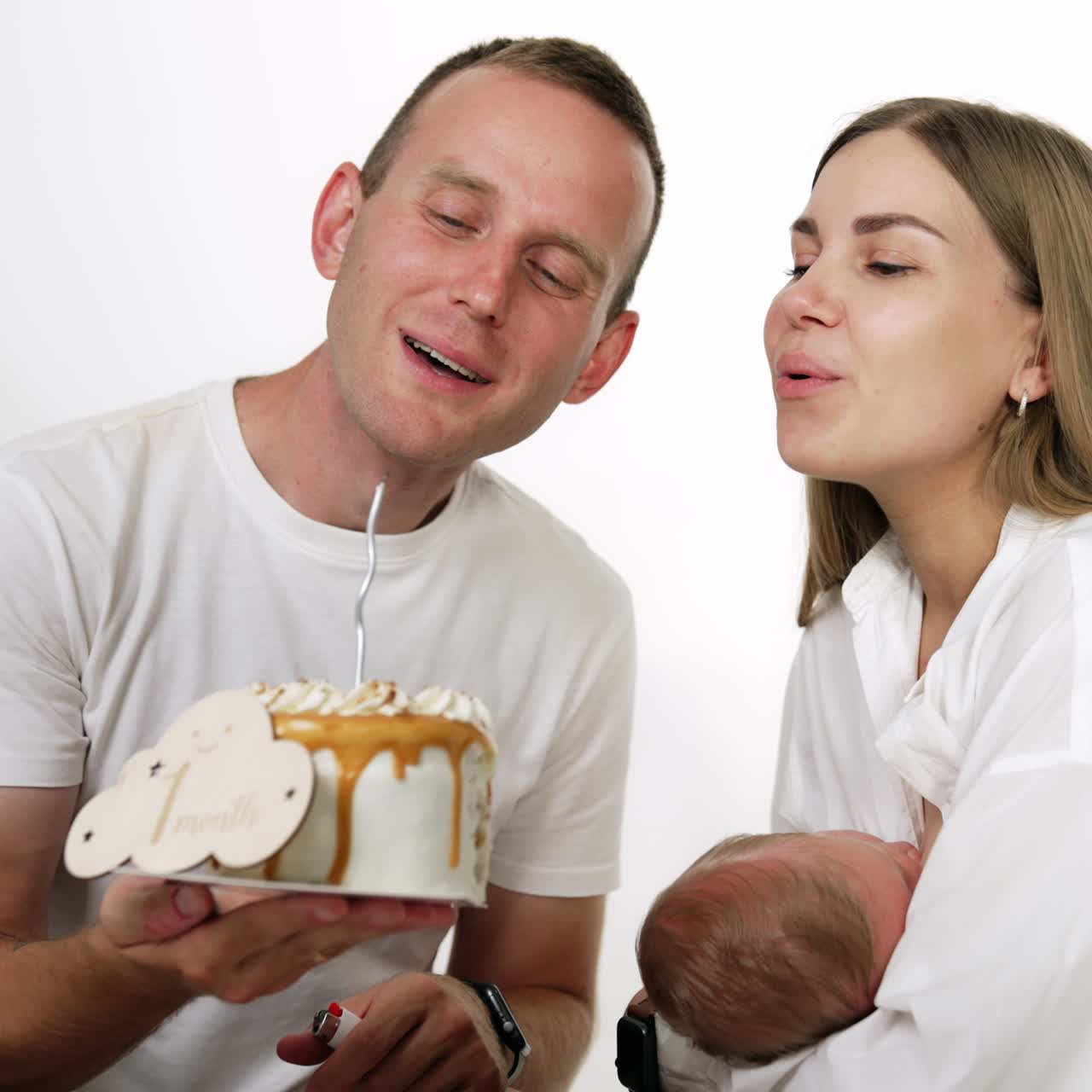 Smiling Caucasian couple with baby blowing a candle on the cake. Family celebrating first month of their infant. White backdrop