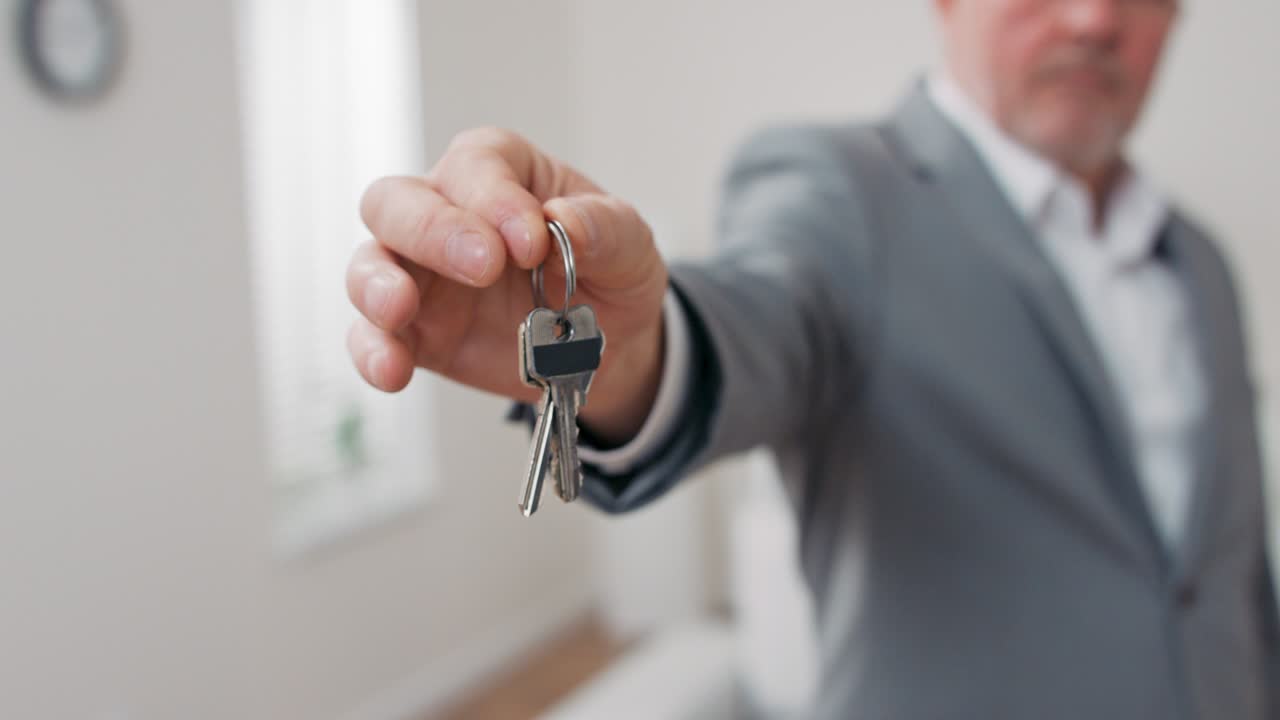 disparo de las llaves del apartamento extendidas en la palma de la mano hacia la cámara, hombre maduro de cabello gris vestido con traje manos sobre las llaves, agente inmobiliario, dueño del apartamento, da espacio para usar