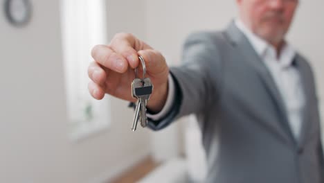 shot of apartment keys extended on palm of hand toward camera, gray-haired mature man dressed in suit hands over keys, real estate agent, owner of apartment, gives room to use