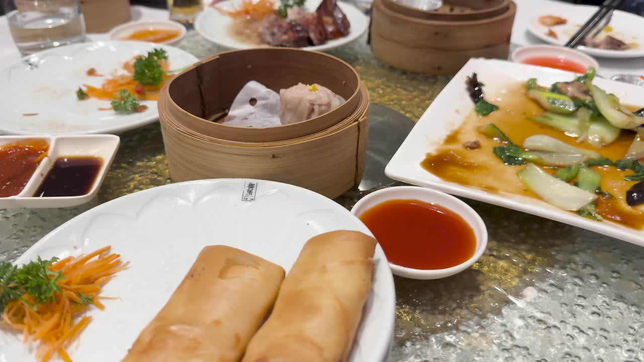 A rotating table displays a variety of dim sum dishes, including spring rolls, vegetables, and sauces, in a bright setting