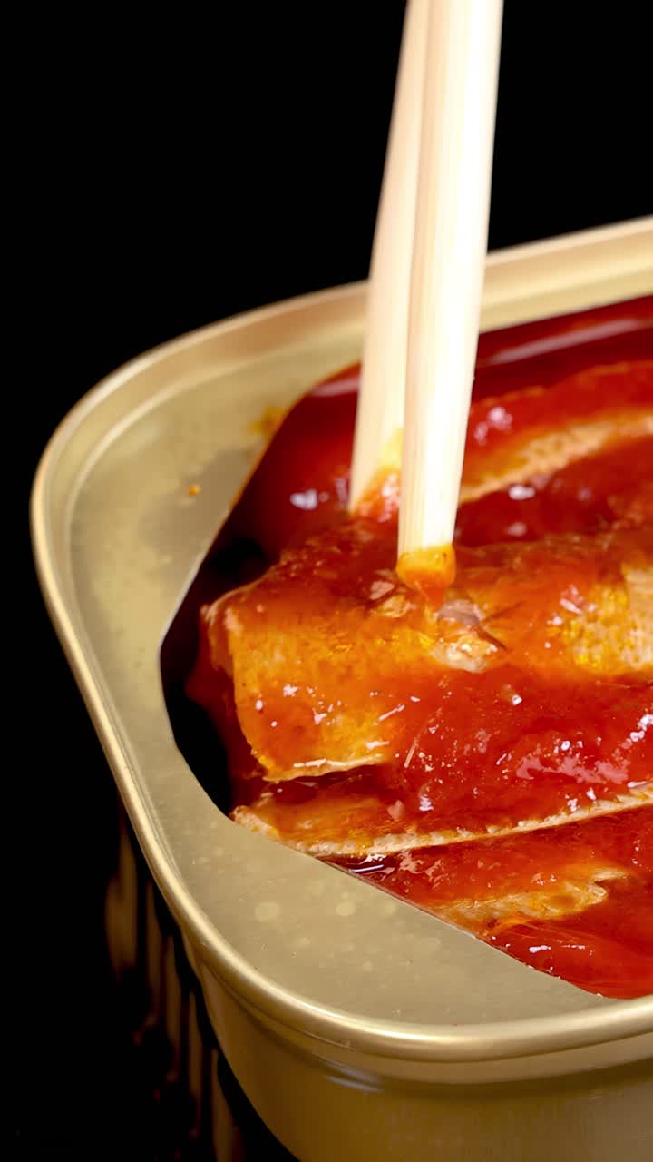 Bacchette afferrano e sollevano una sardina da una lattina aperta piena di salsa di pomodoro, sotto una brillante illuminazione da studio e su uno sfondo nero