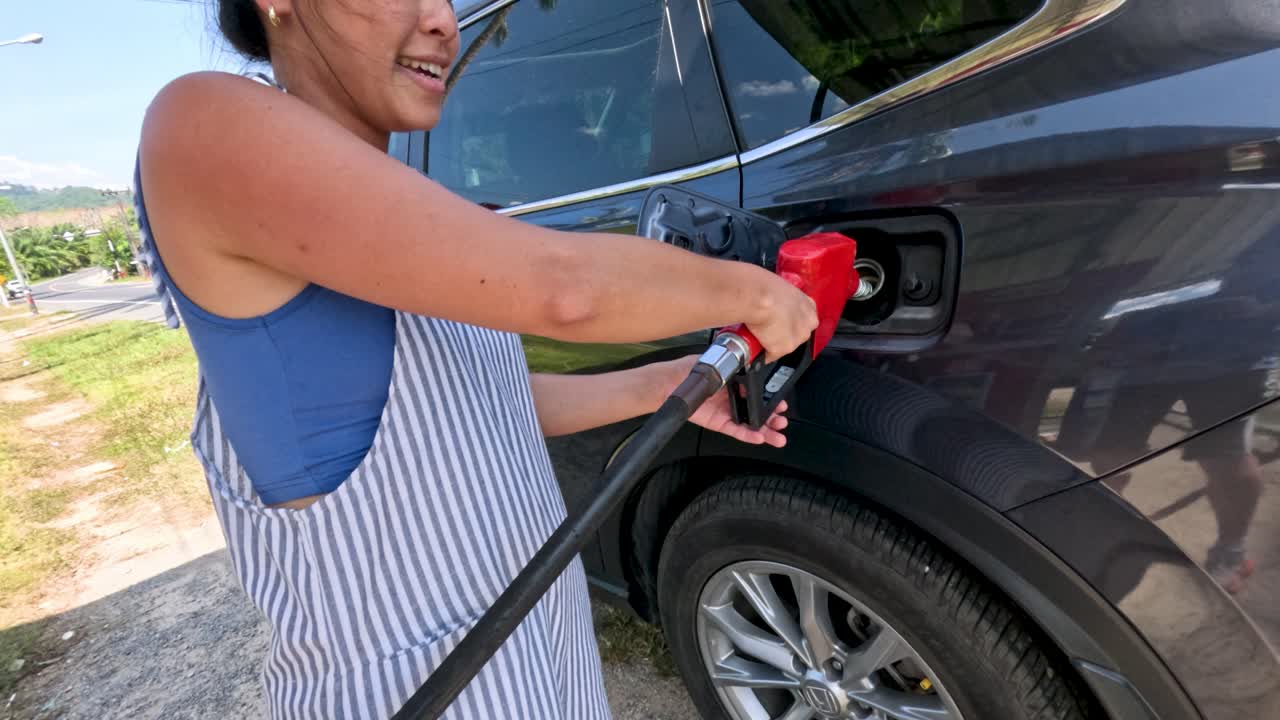 Smiling woman fills black SUV with fuel outdoors, bright daylight, handheld camera, casual atmosphere