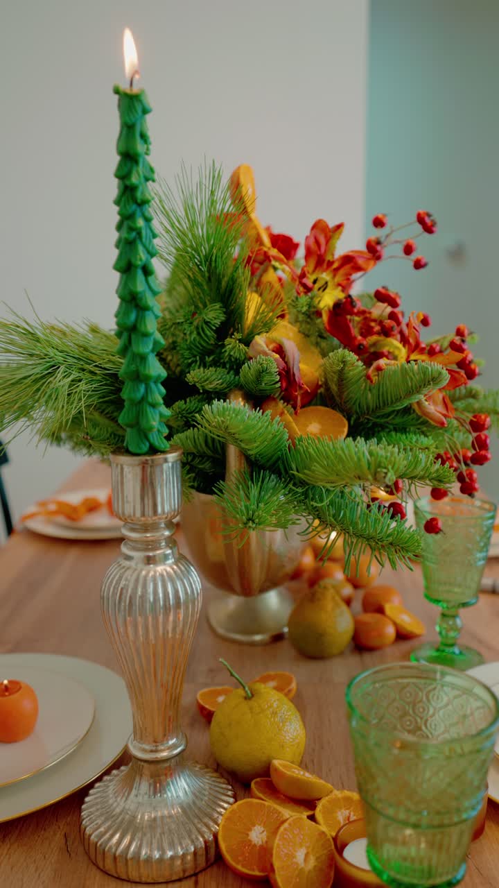 An elegant New Year's Eve tablescape featuring a vibrant centerpiece of fresh pine, dried citrus, and mandarin oranges. Green taper candles illuminate the festive holiday dinner setting