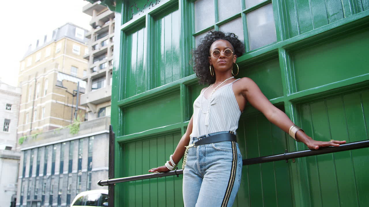 Trendy young black woman waring round sunglasses, camisole and jeans leaning on a handrail in a city street, low angle
