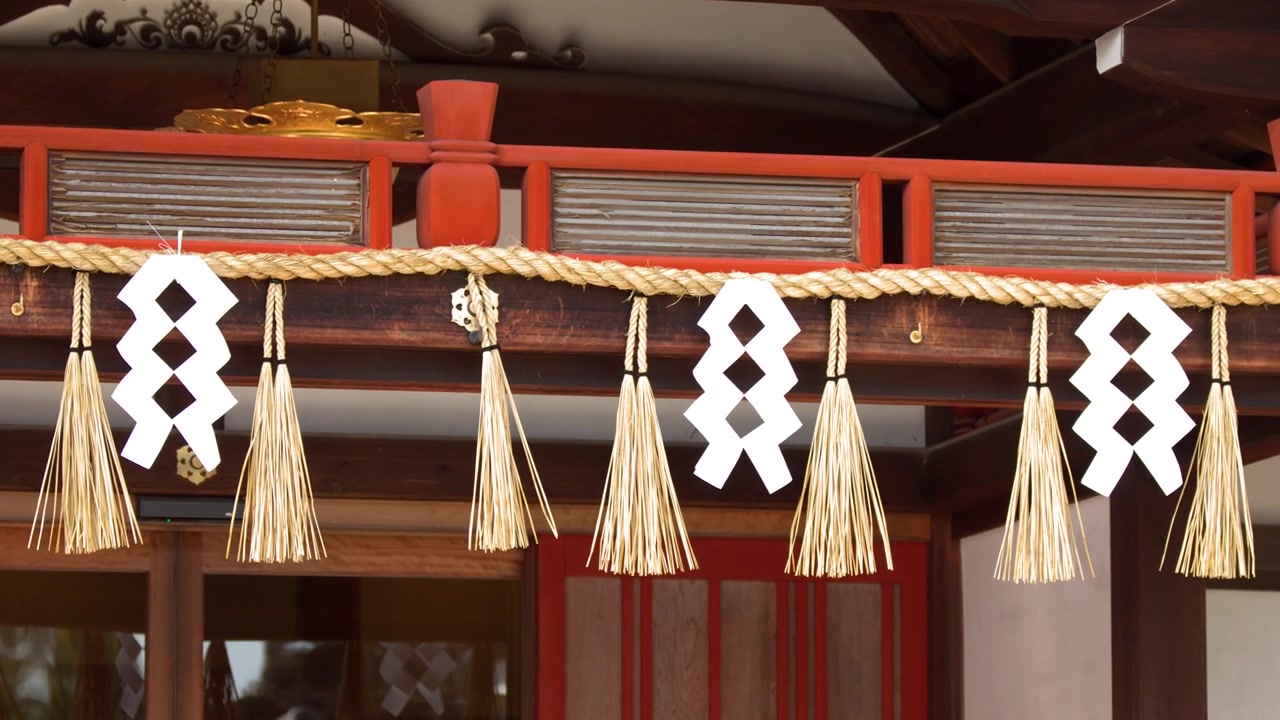 Static shot of sacred straw rope and paper tassels hanging from red wooden temple architecture.