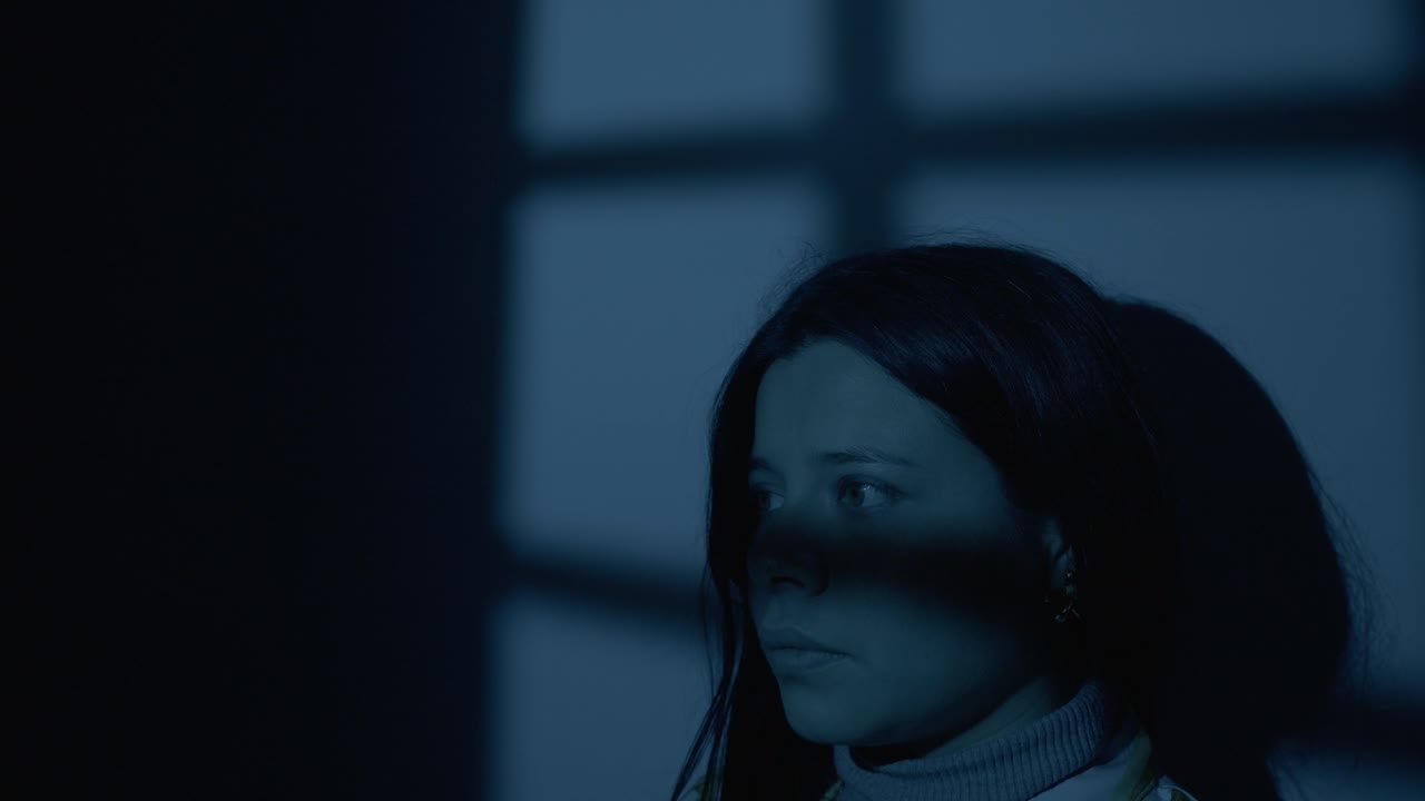 A woman looks thoughtful as she stands in front of her bedroom window after nightfall